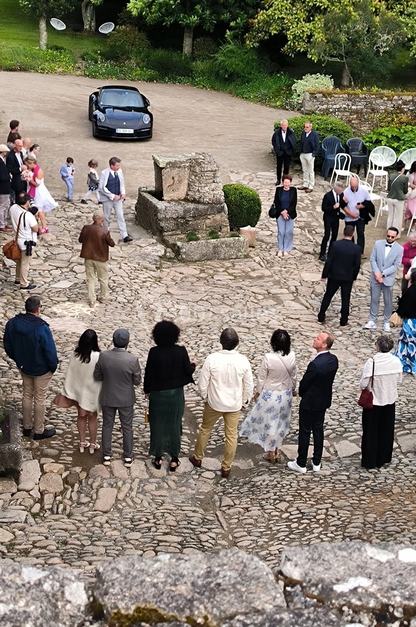 Groupe de personnes rassemblées en plein air sur une place pavée, avec des chaises et une voiture en arrière-plan.