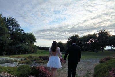 Vue aérienne d'un manoir en pierre entouré de champs et de végétation, avec un village et la mer à l'horizon.