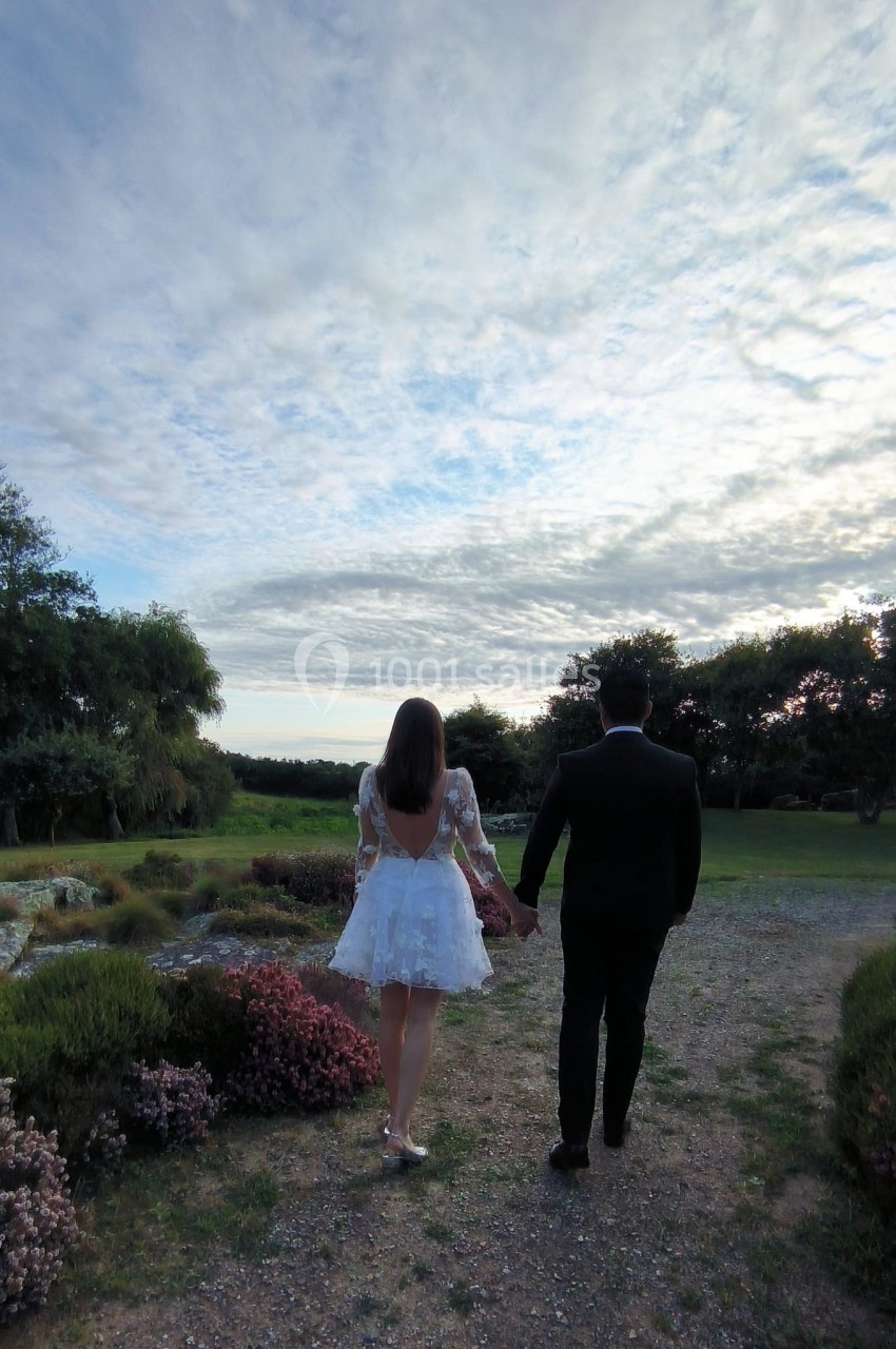 Un couple marchant main dans la main sur un chemin entouré de végétation, sous un ciel partiellement nuageux.