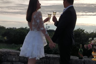 Vue aérienne d'un manoir en pierre entouré de champs et de végétation, avec un village et la mer à l'horizon.