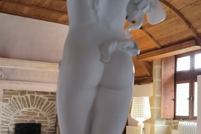 Vue aérienne d'un manoir en pierre entouré de champs et de végétation, avec un village et la mer à l'horizon.