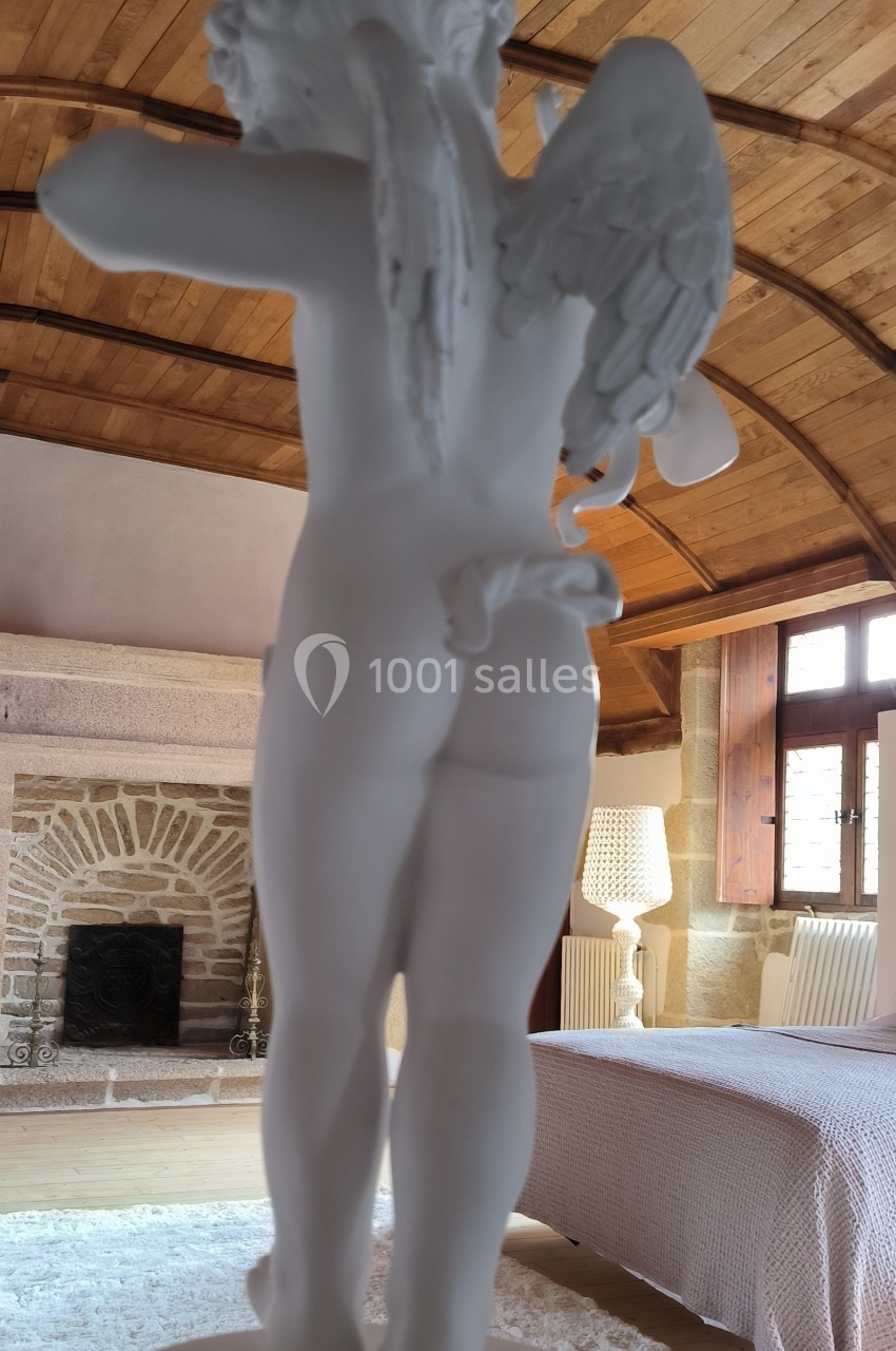 Statue blanche d'un ange vu de dos, exposée dans une pièce avec un plafond en bois et une cheminée en pierre.
