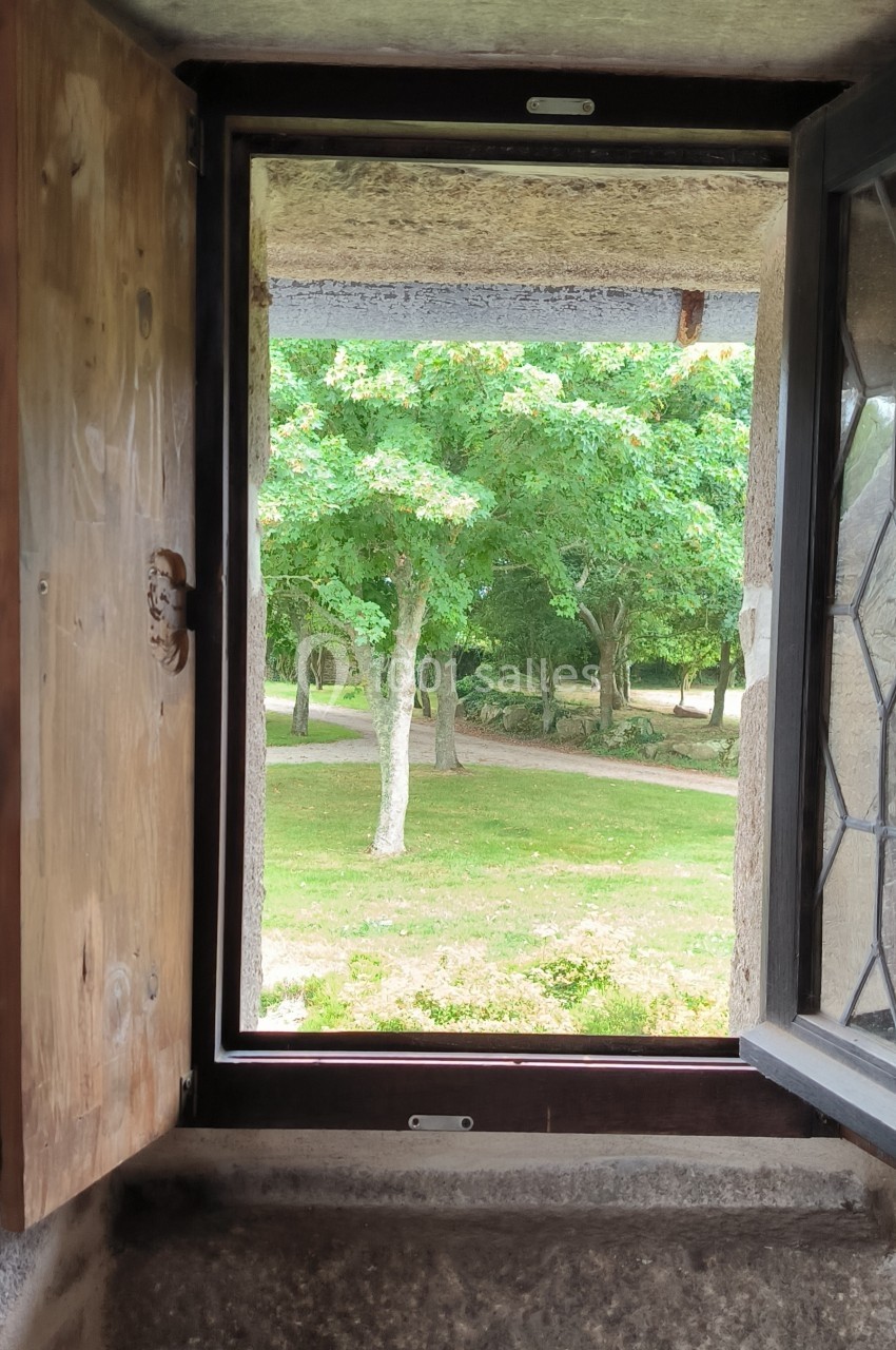 Vue sur un parc verdoyant à travers une fenêtre en bois et en pierre, partiellement ouverte.