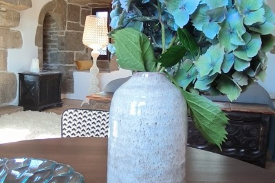 Vue aérienne d'un manoir en pierre entouré de champs et de végétation, avec un village et la mer à l'horizon.