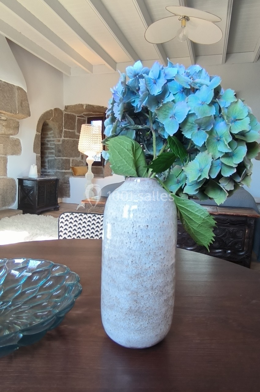 Vase en céramique blanche contenant des hortensias bleus, posé sur une table en bois dans un intérieur lumineux.