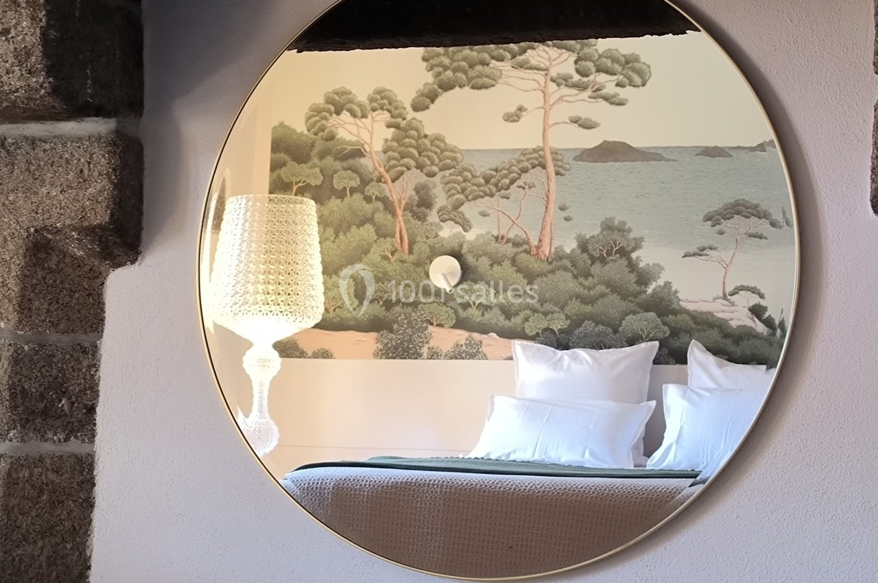 Reflet d'une chambre avec un lit, une lampe et une fresque murale représentant un paysage côtier, dans un miroir rond.