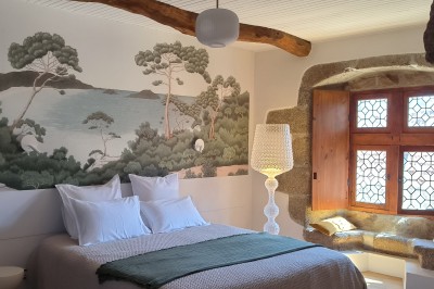 Vue aérienne d'un manoir en pierre entouré de champs et de végétation, avec un village et la mer à l'horizon.