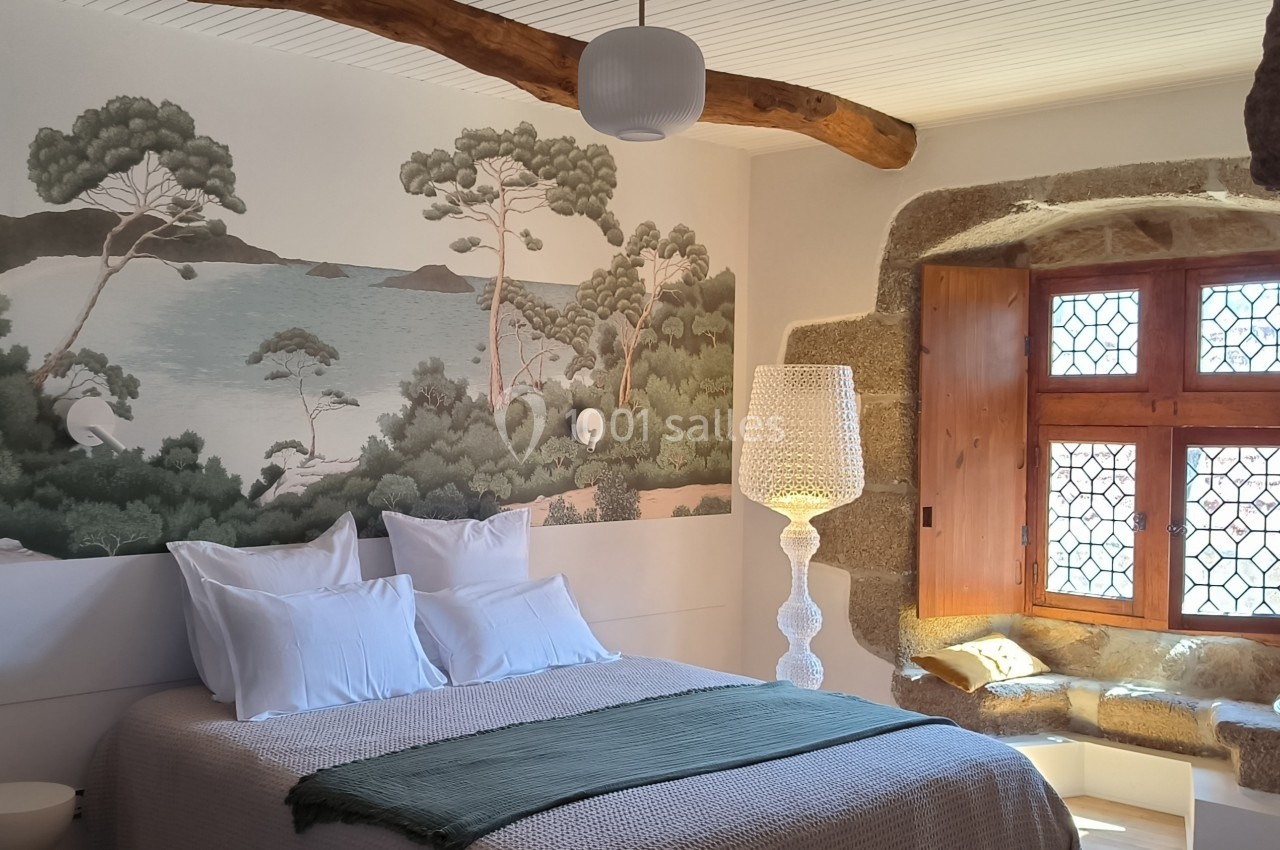 Chambre lumineuse avec lit double, fresque murale représentant un paysage côtier, fenêtre en pierre et coin lecture.