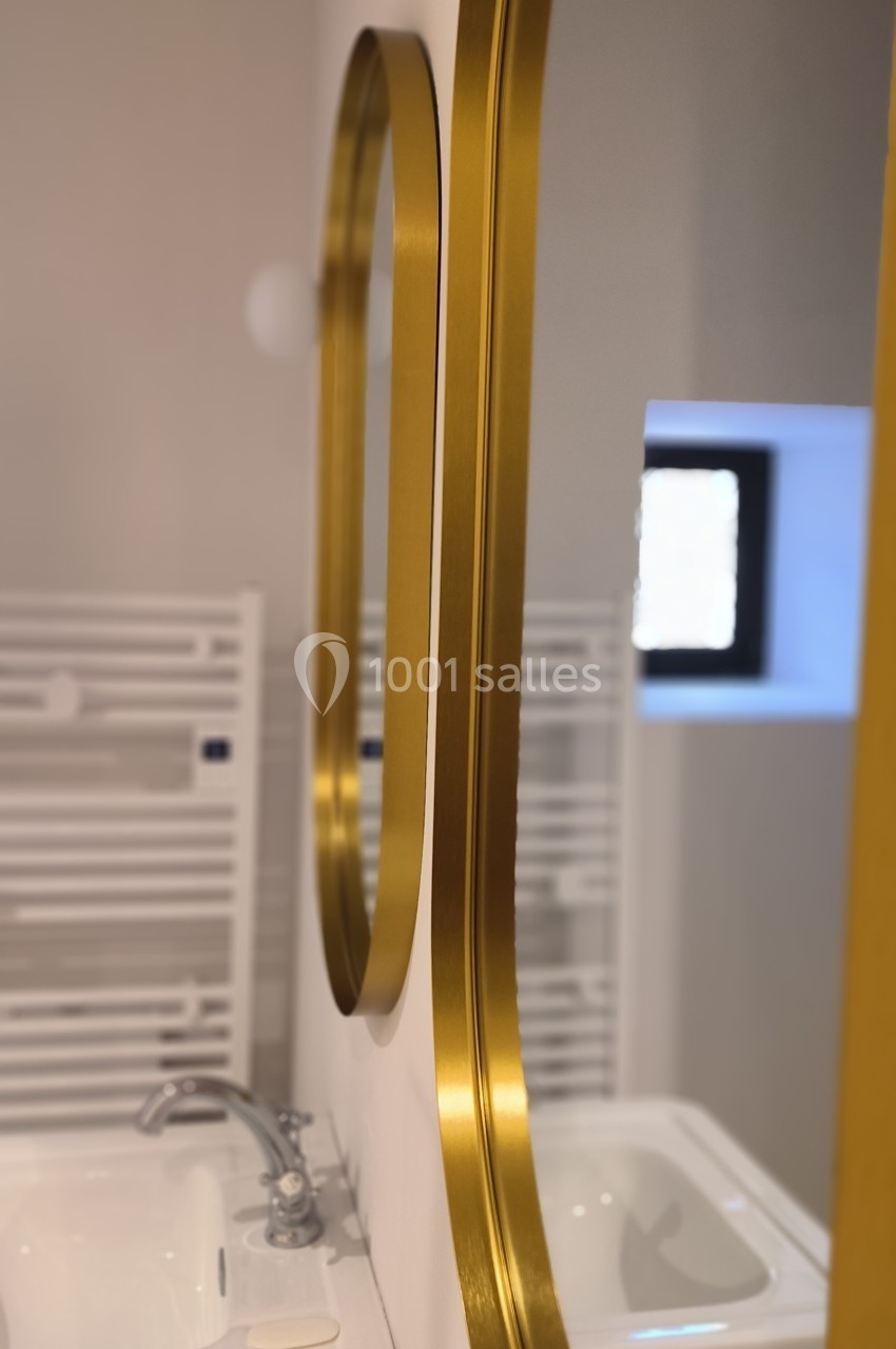 Miroir doré ovale reflétant un lavabo blanc et un sèche-serviettes dans une salle de bain lumineuse.