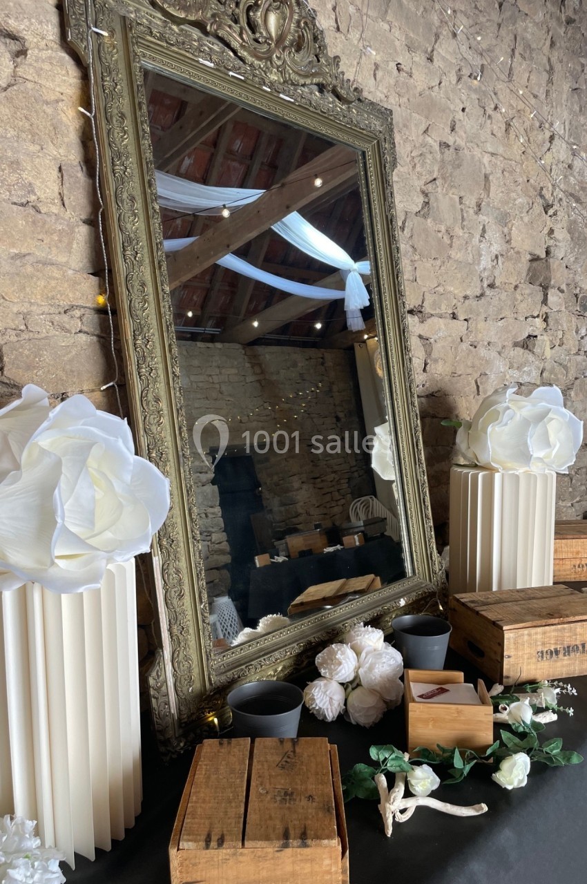 Grand miroir doré encadré, posé contre un mur en pierre, entouré de décorations florales et de caisses en bois.