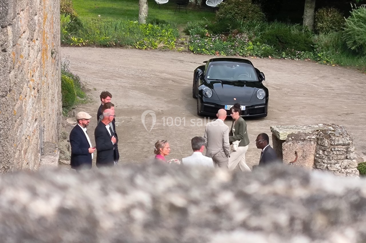 Un groupe de personnes discute dans une cour pavée, avec une voiture noire garée en arrière-plan.