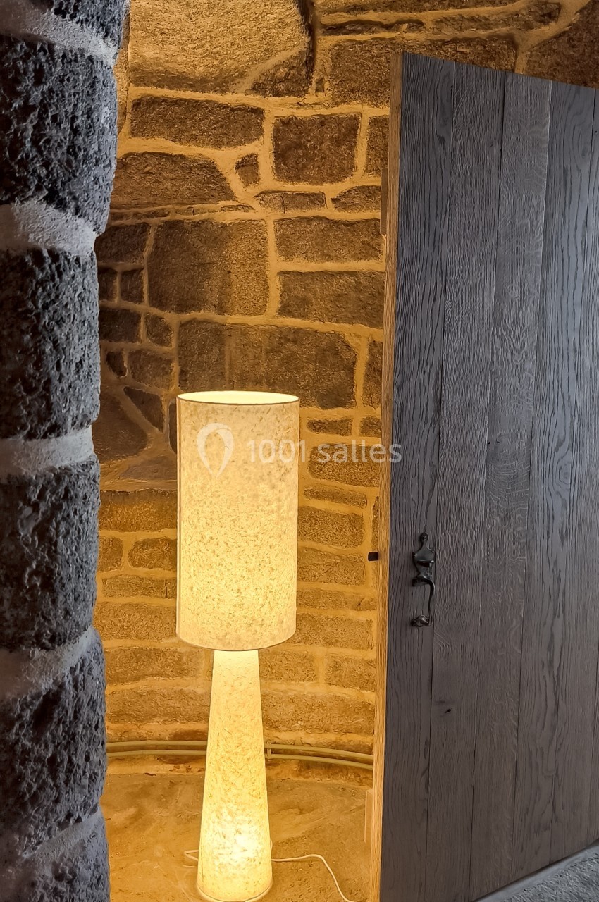 Lampe allumée posée au sol près d'un mur en pierre et d'une porte en bois entrouverte.