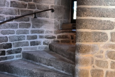 Vue aérienne d'un manoir en pierre entouré de champs et de végétation, avec un village et la mer à l'horizon.