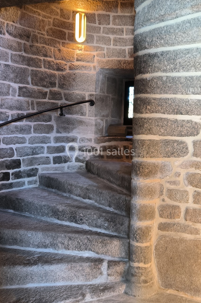 Escalier en pierre courbé avec rampe en métal, éclairé par une applique murale et une petite fenêtre.