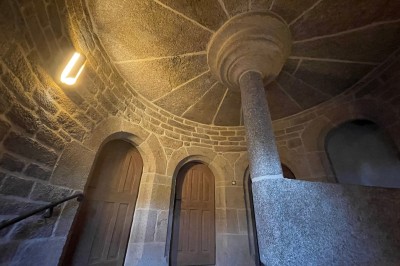 Vue aérienne d'un manoir en pierre entouré de champs et de végétation, avec un village et la mer à l'horizon.
