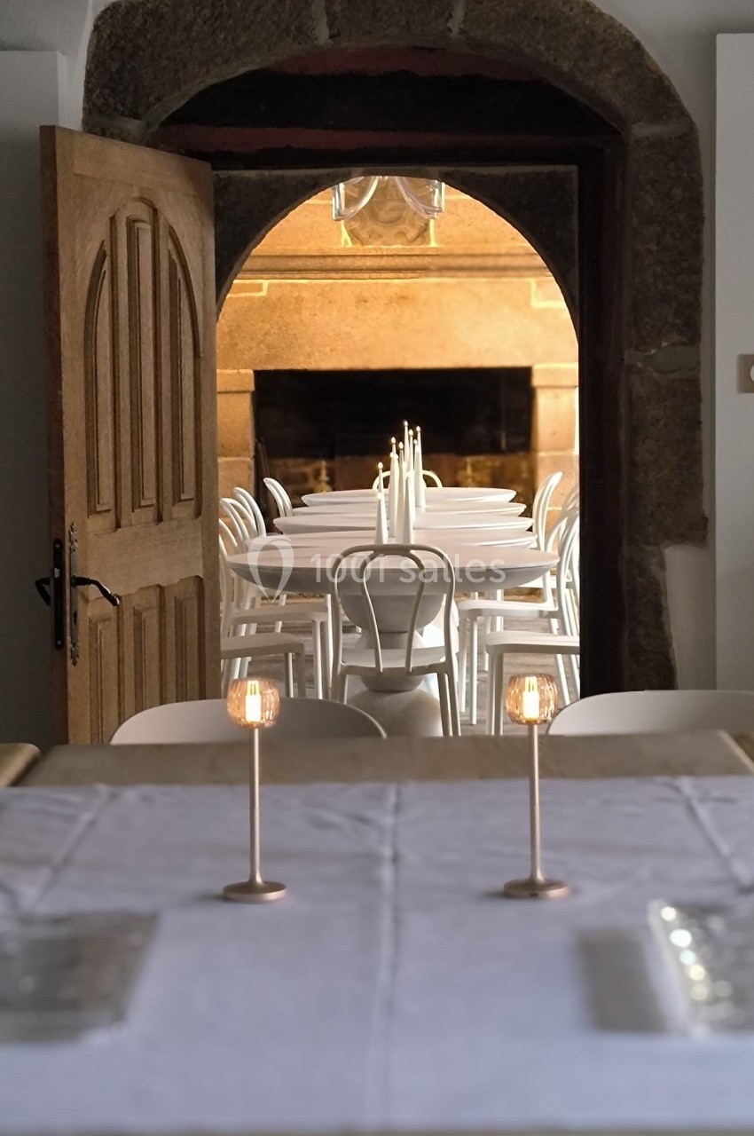 Vue d'une salle à manger avec des tables dressées, des chaises blanches et une porte en bois ouverte sur un autre espace.