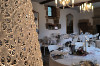 Vue aérienne d'un manoir en pierre entouré de champs et de végétation, avec un village et la mer à l'horizon.