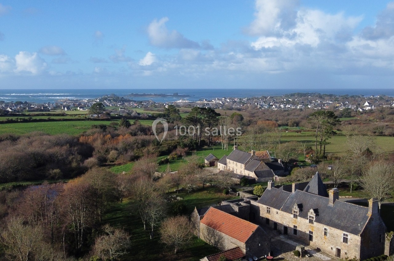 Vue aérienne d'un manoir en pierre entouré de champs et de végétation, avec un village et la mer à l'horizon.
