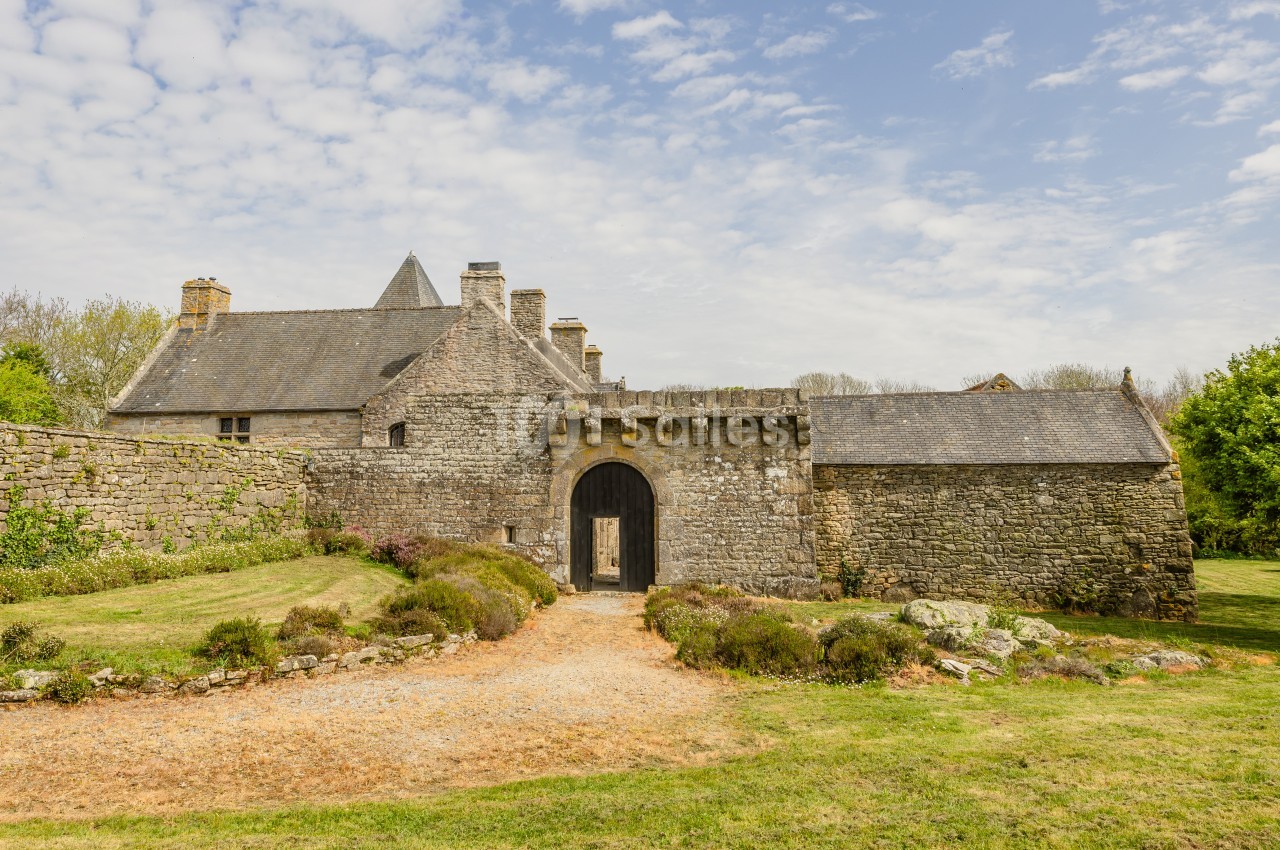 Façade en pierre d'un manoir avec portail cintré en bois sombre, toit d'ardoise et tourelle conique, devant une allée de gravier et une pelouse.