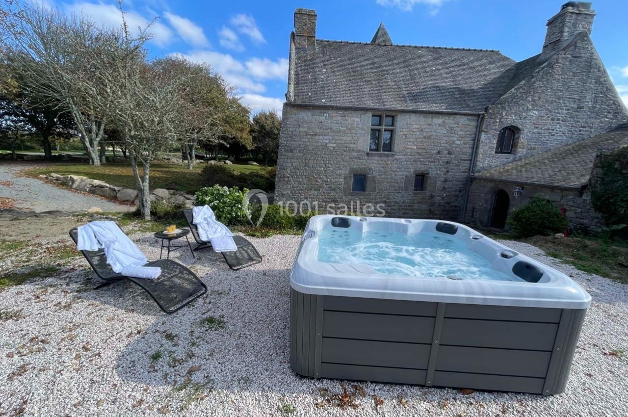 Jacuzzi extérieur bouillonnant devant une maison en pierre, entouré de chaises longues et d'arbres sous un ciel dégagé.