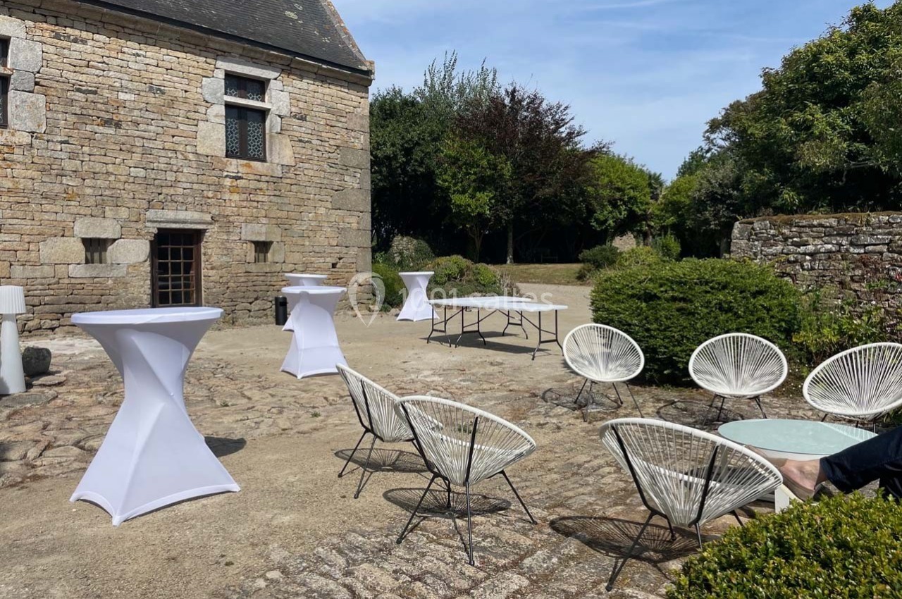 Cour extérieure en pierre avec des tables hautes couvertes de nappes blanches et des fauteuils disposés autour de petites…