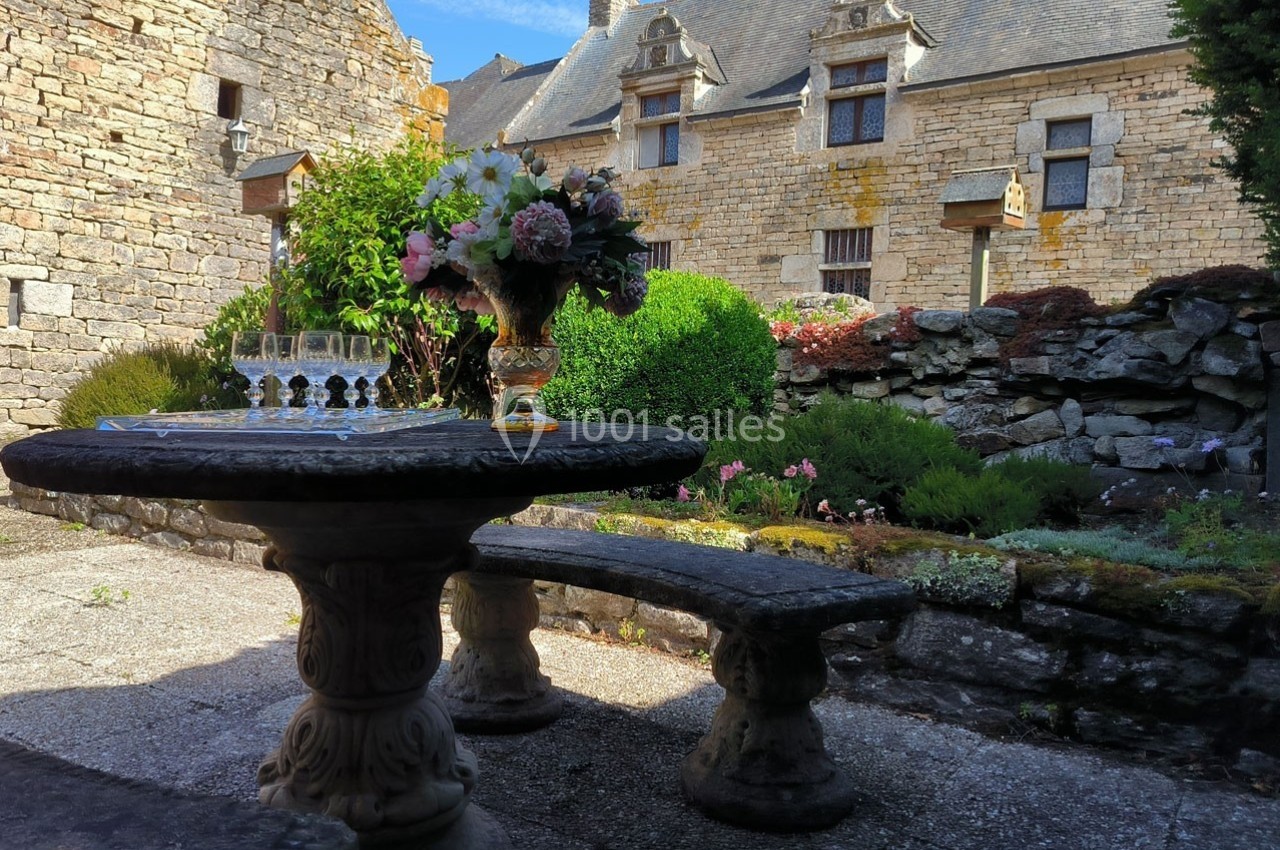 Table en pierre avec un bouquet de fleurs et des verres, située dans une cour pavée entourée de bâtiments en pierre.