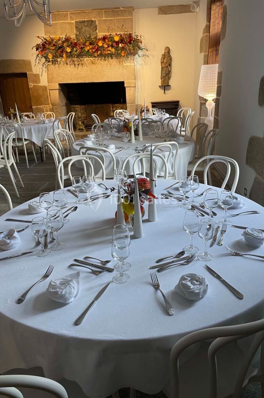 Salle de réception avec tables rondes dressées, nappes blanches, chaises blanches et décoration florale au mur.