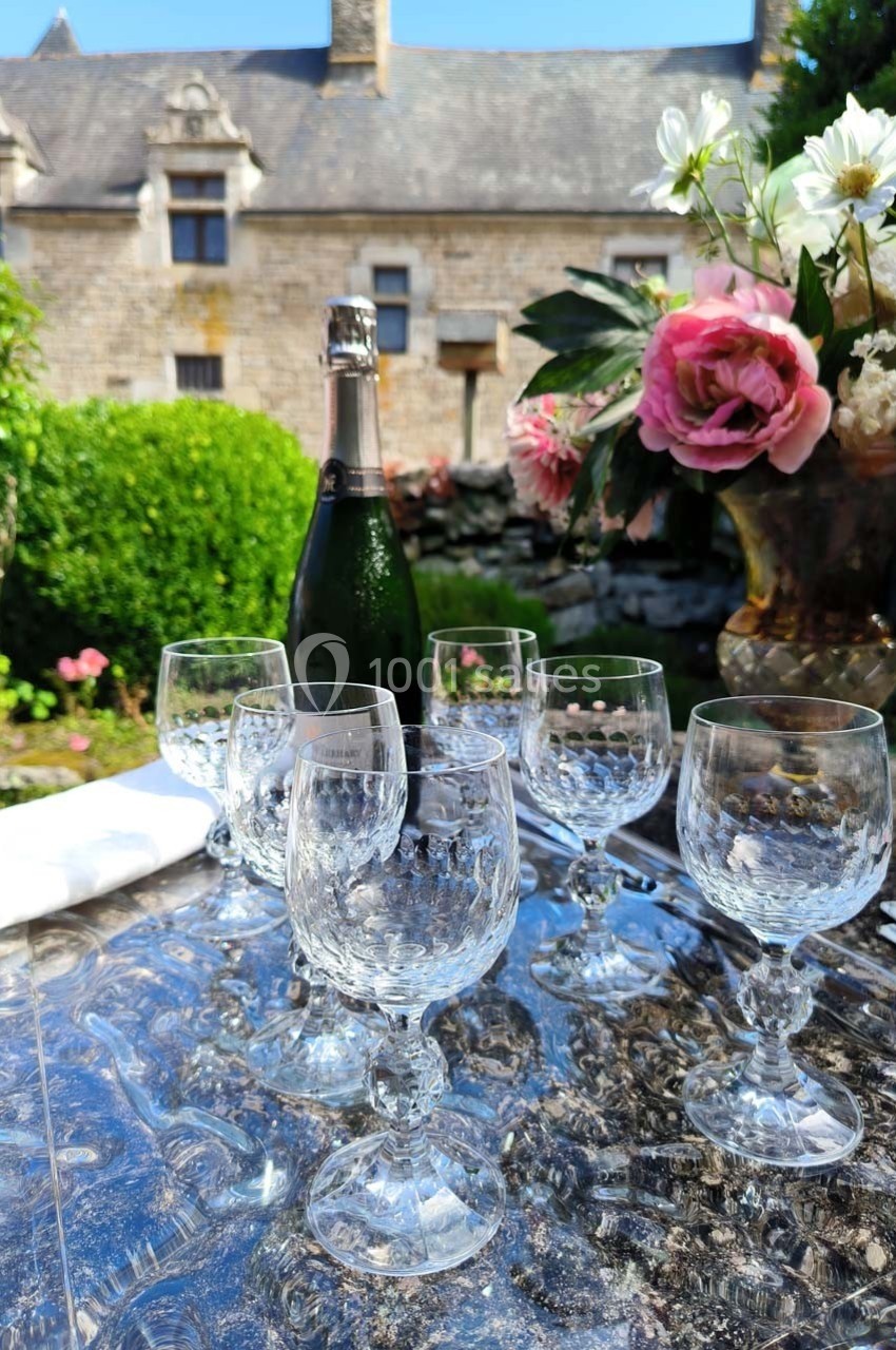 Bouteille de champagne et verres en cristal disposés sur une table en extérieur, avec un jardin et un bâtiment en arrière…