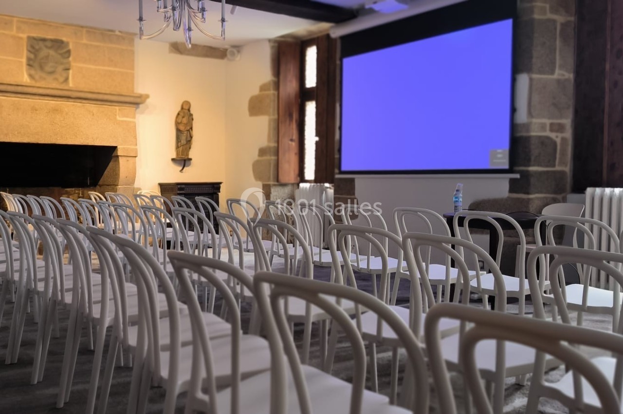 Salle de conférence avec rangées de chaises blanches, écran de projection allumé et cheminée en pierre.