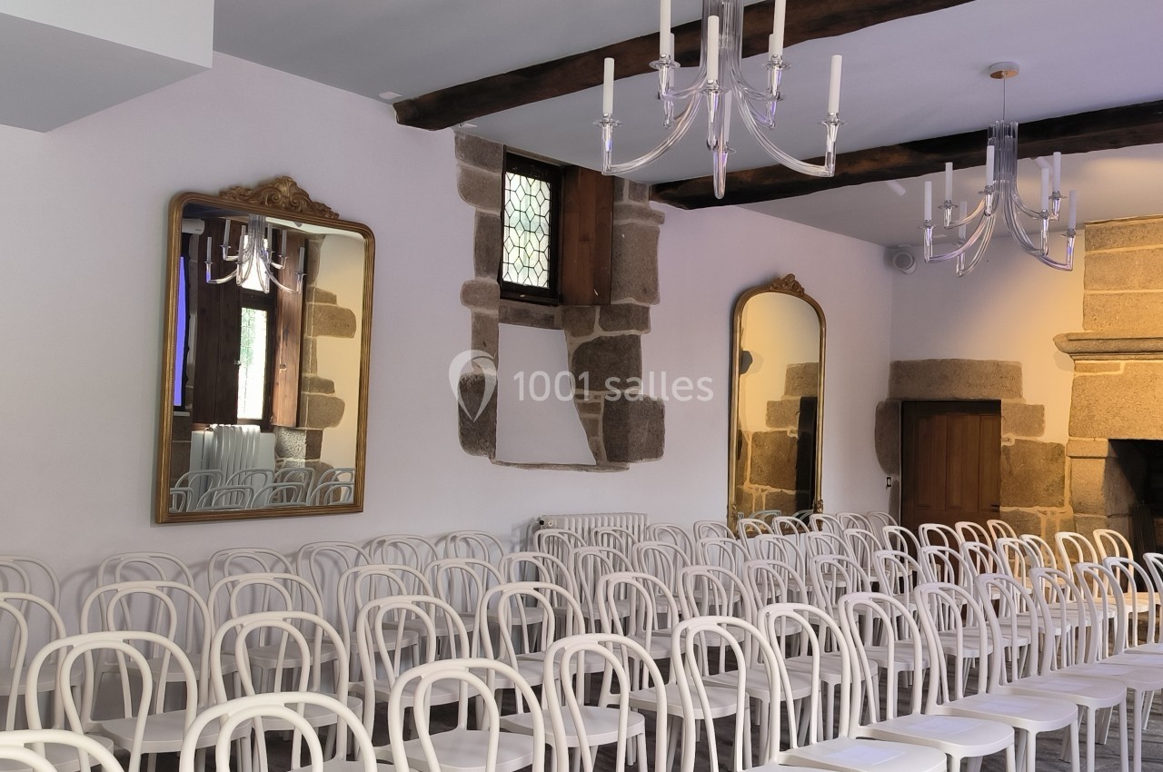 Salle avec des chaises blanches alignées, murs en pierre, miroirs, lustres et une grande cheminée en arrière-plan.