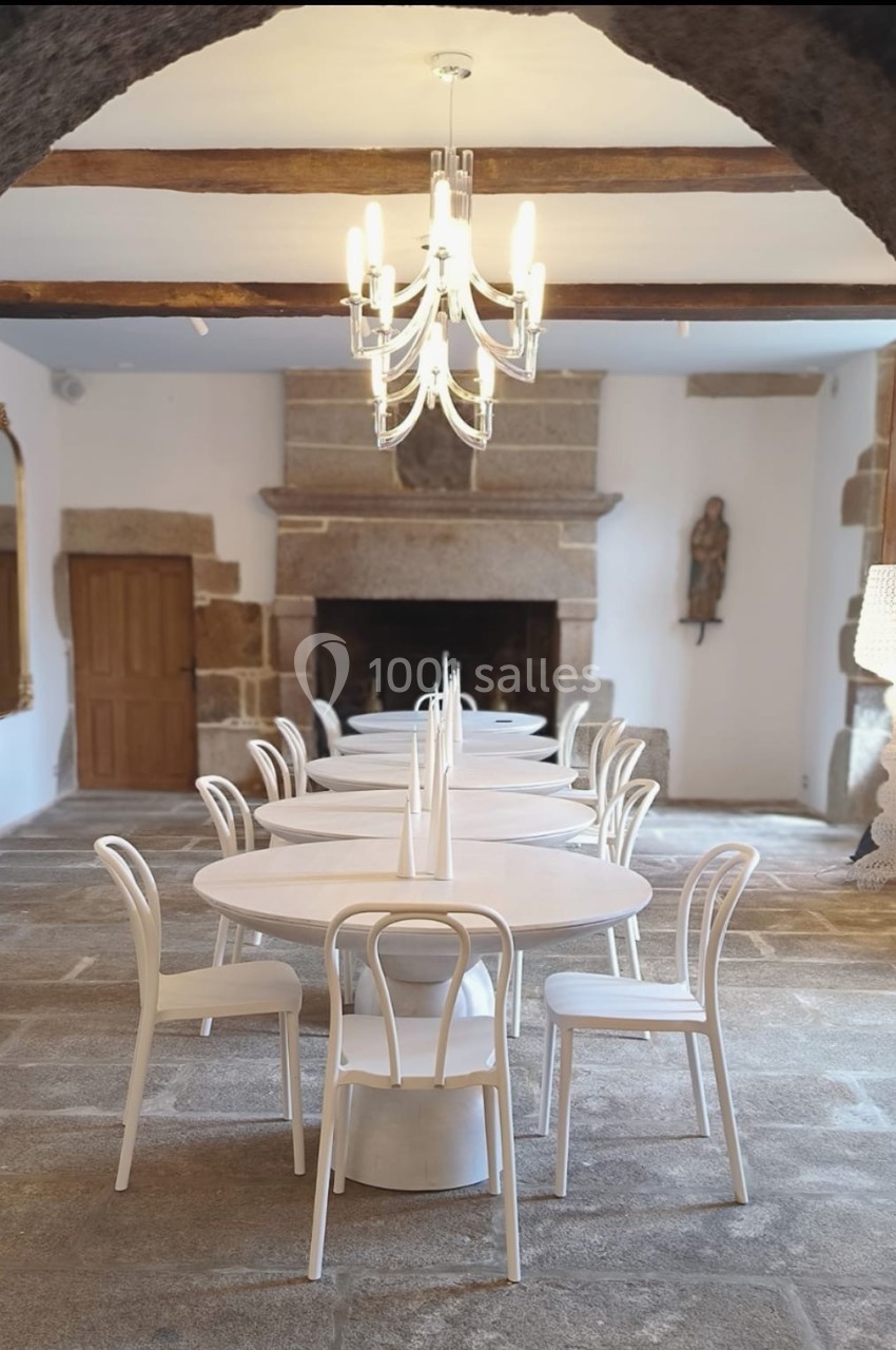 Salle lumineuse avec tables rondes blanches, chaises assorties, lustre moderne et cheminée en pierre.