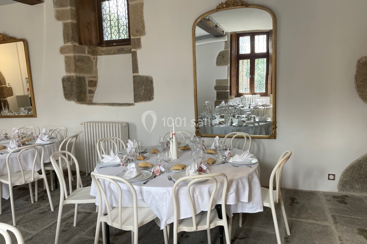 Salle de réception avec tables dressées, nappes blanches, chaises blanches et miroirs encadrés sur des murs en pierre.
