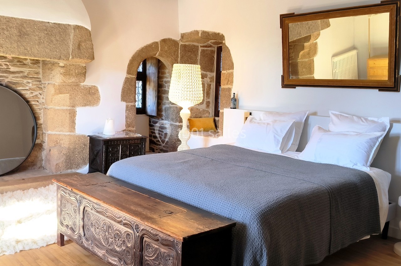 Chambre lumineuse avec lit double, murs en pierre, miroir rond, coffre en bois et lampe suspendue.