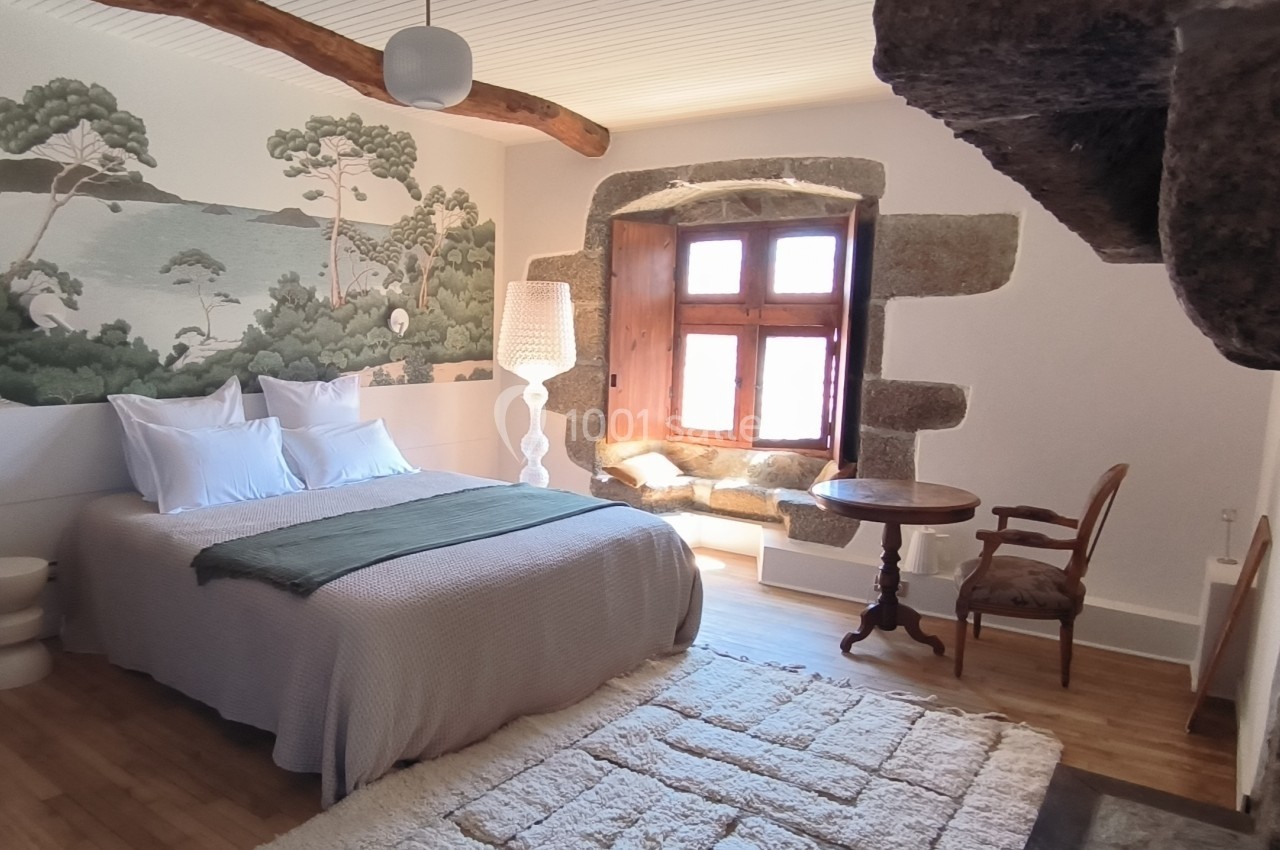 Chambre lumineuse avec lit double, mur décoré d'une fresque nature, poutres apparentes et fenêtre en pierre.