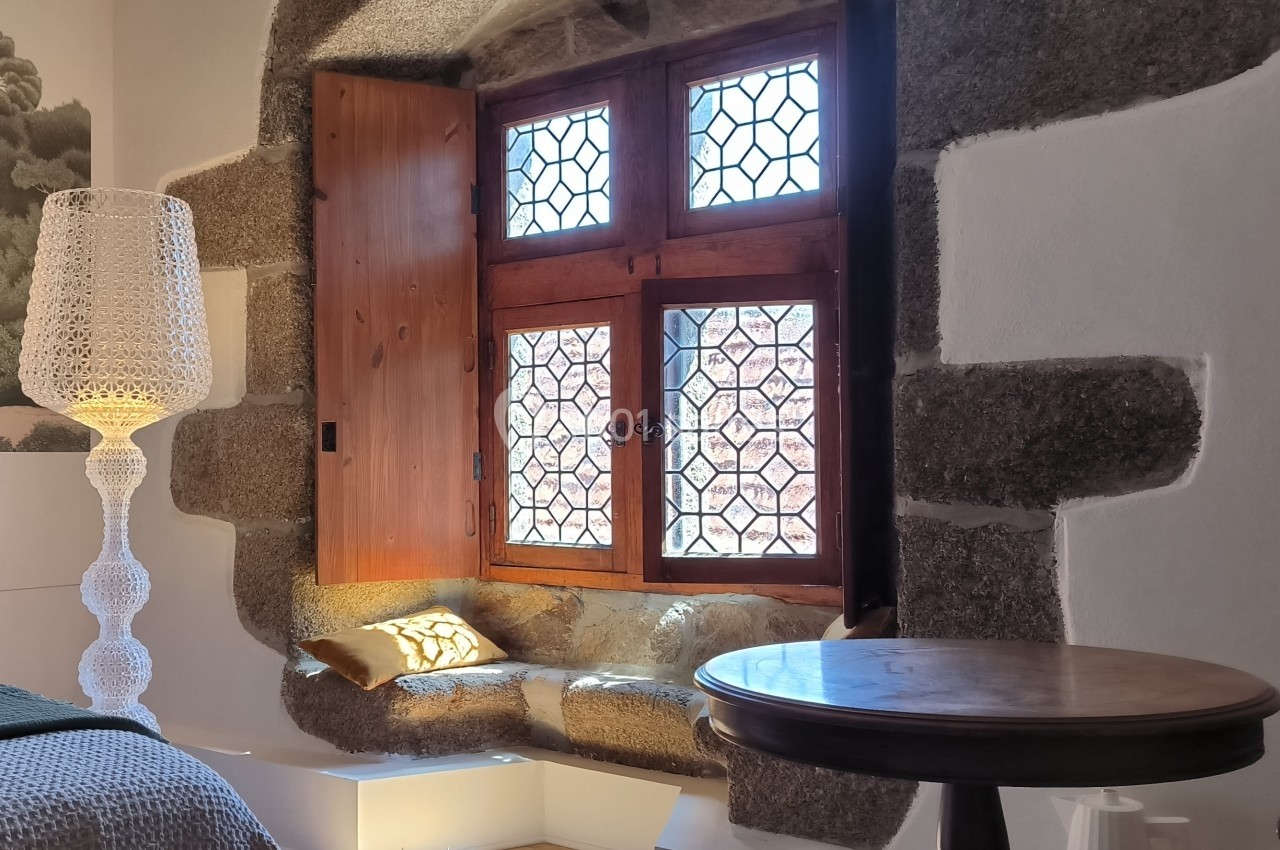 Fenêtre ancienne avec vitraux, encadrée de pierre, un coussin sur l'assise et une lampe design à côté.