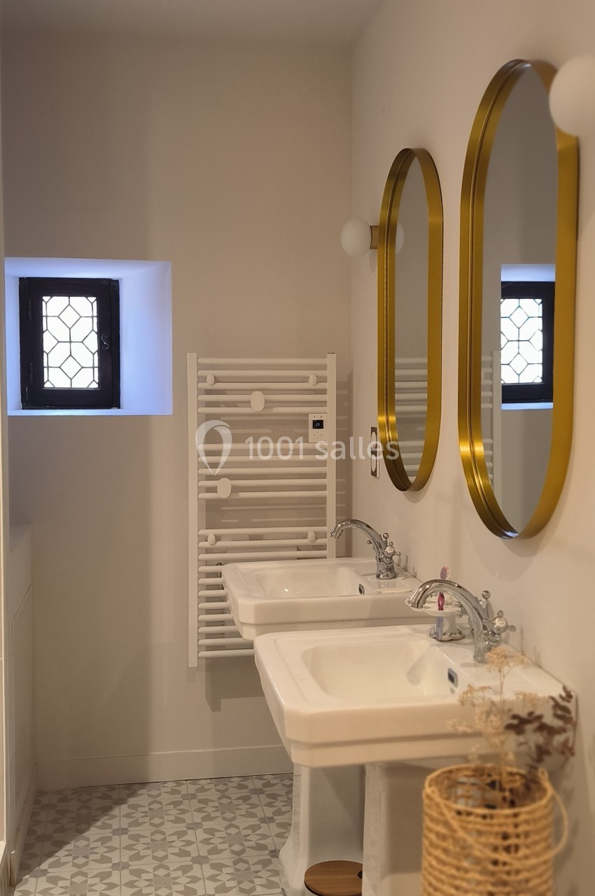 Salle de bain lumineuse avec deux lavabos, miroirs dorés, radiateur blanc et petite fenêtre à grille.
