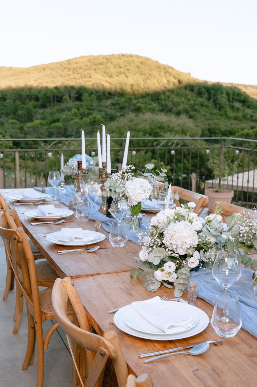 Table en bois dressée avec vaisselle blanche, bougies et compositions florales, sur une terrasse avec vue sur des collines…