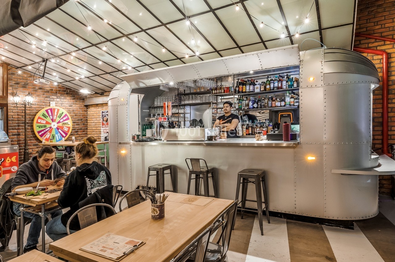 Intérieur d'un restaurant avec un bar en métal, des tables en bois, et un serveur derrière le comptoir.
