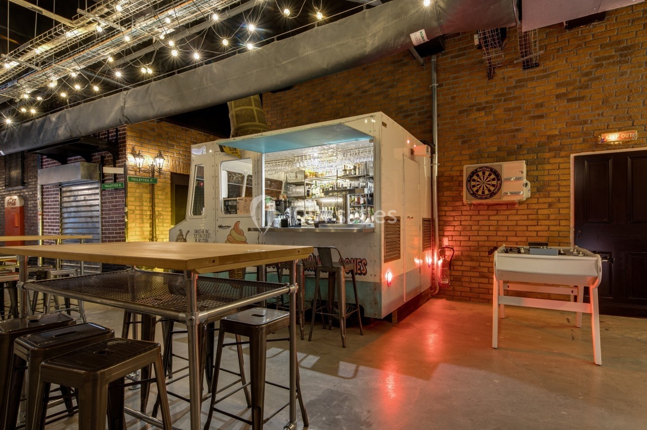 Bar aménagé dans un conteneur, entouré de tables hautes, baby-foot et décoré de guirlandes lumineuses suspendues.