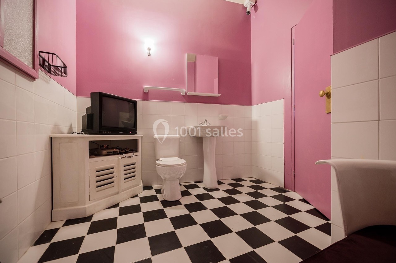 Salle de bain avec murs roses, carrelage noir et blanc, lavabo, toilettes, et meuble avec une télévision.
