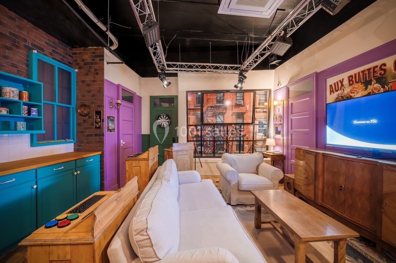 Salon coloré avec canapé blanc, meubles en bois, décorations murales et écran allumé dans un cadre chaleureux.