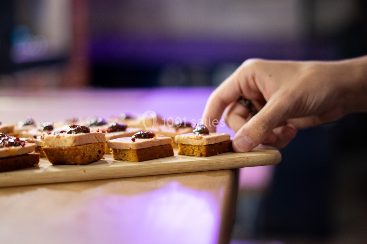 Main prenant un canapé garni de pâté et de confiture sur un plateau en bois, dans un environnement flou.