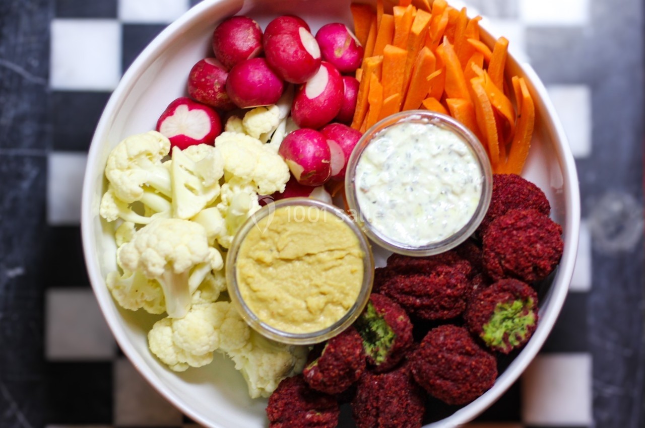 Assiette composée de légumes crus, falafels rouges et deux sauces, présentée sur une table à motif damier.