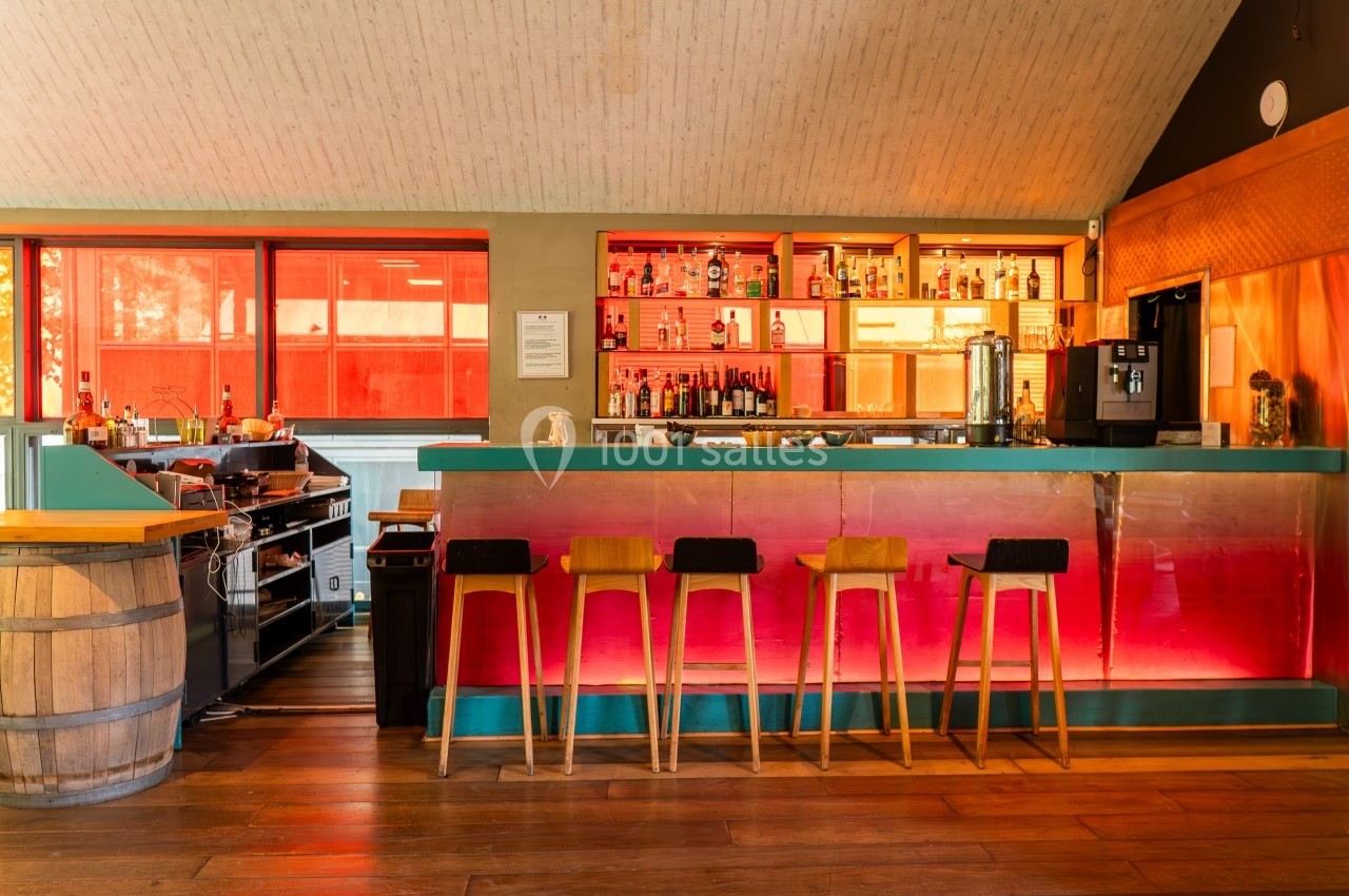 Bar moderne avec comptoir en métal, tabourets en bois, étagères éclairées contenant des bouteilles et fenêtres rouges en…