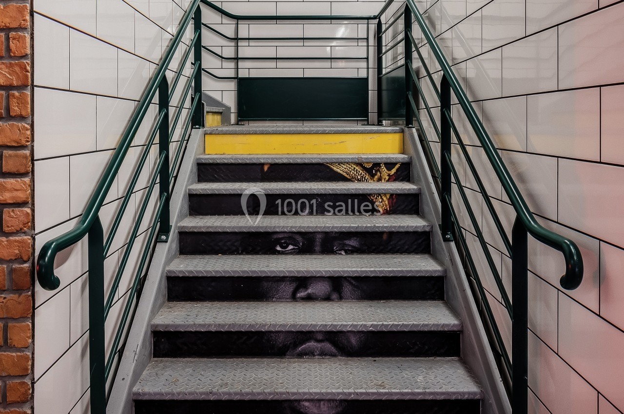 Escalier de métro avec une fresque représentant un visage sur les marches, entouré de murs carrelés blancs.