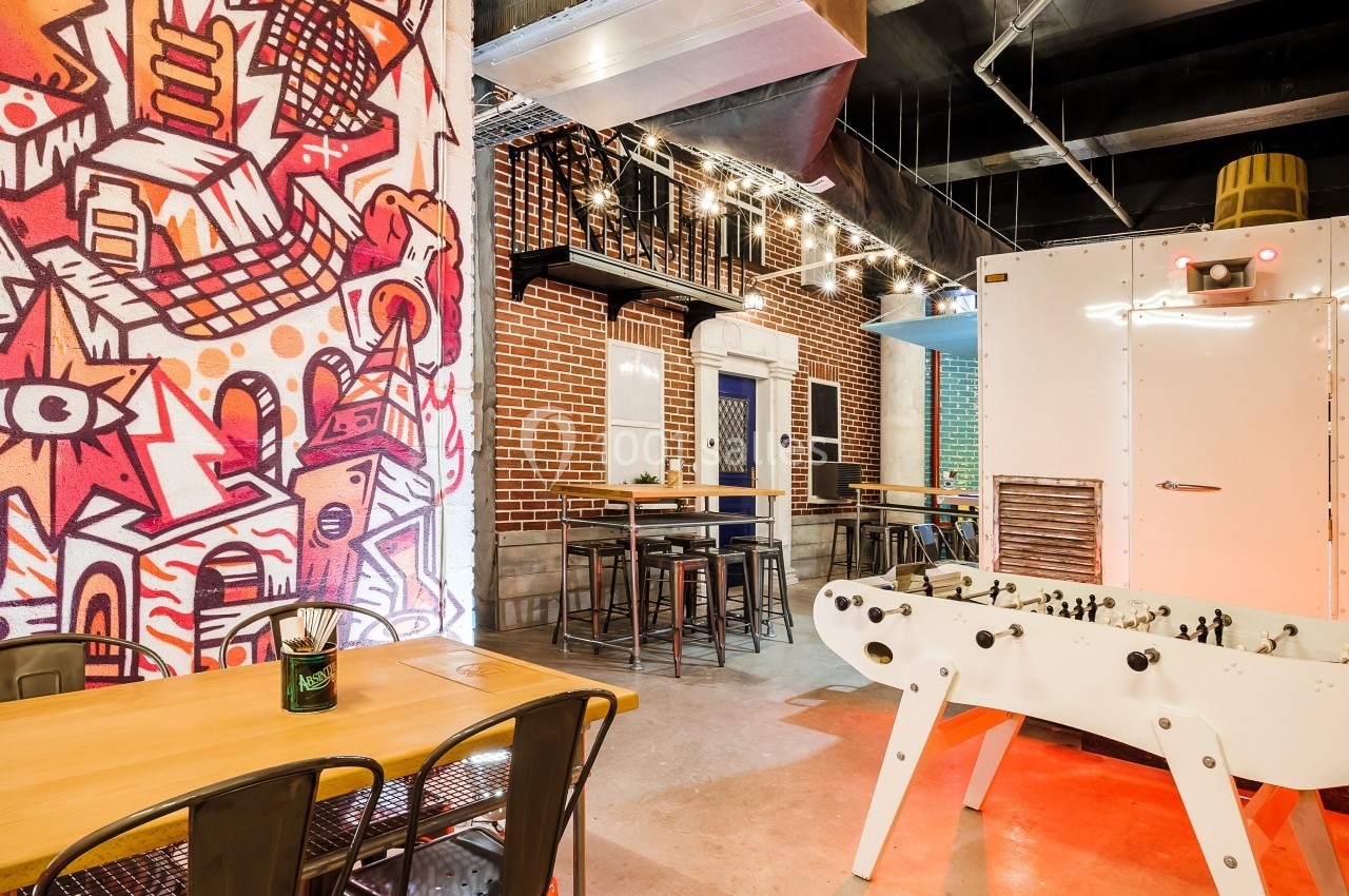 Salle de détente avec baby-foot, tables en bois, chaises métalliques et mur décoré d'une fresque colorée.
