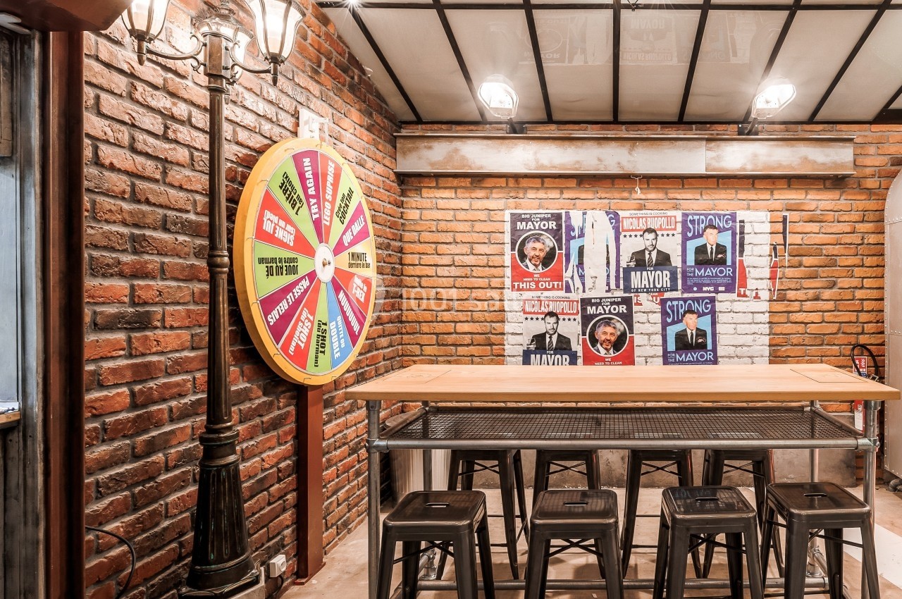 Salle au style industriel avec murs en briques, table haute, tabourets métalliques et une roue colorée accrochée au mur.