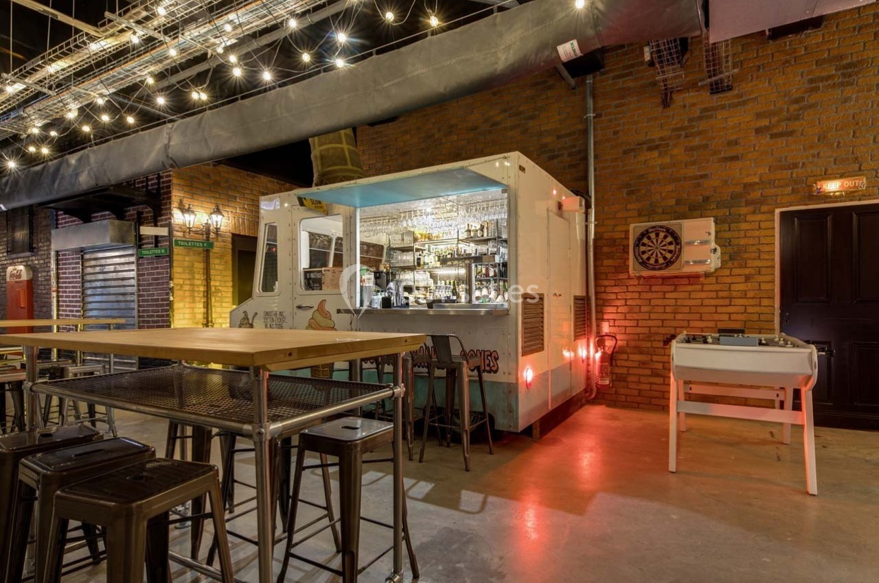 Bar aménagé dans un conteneur blanc, entouré de tables hautes, avec des guirlandes lumineuses suspendues au plafond.