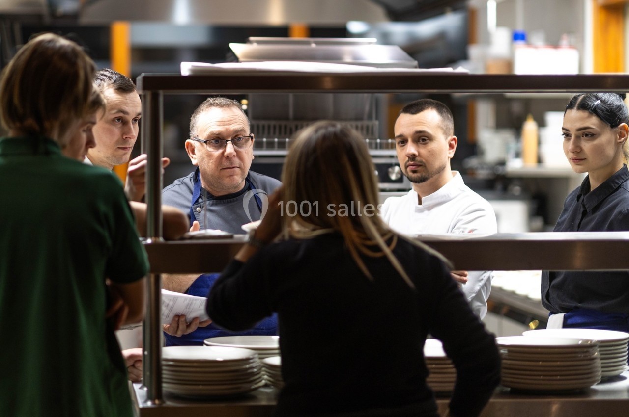 Un groupe de cuisiniers et une femme discutent dans une cuisine professionnelle près d'un passe-plat.