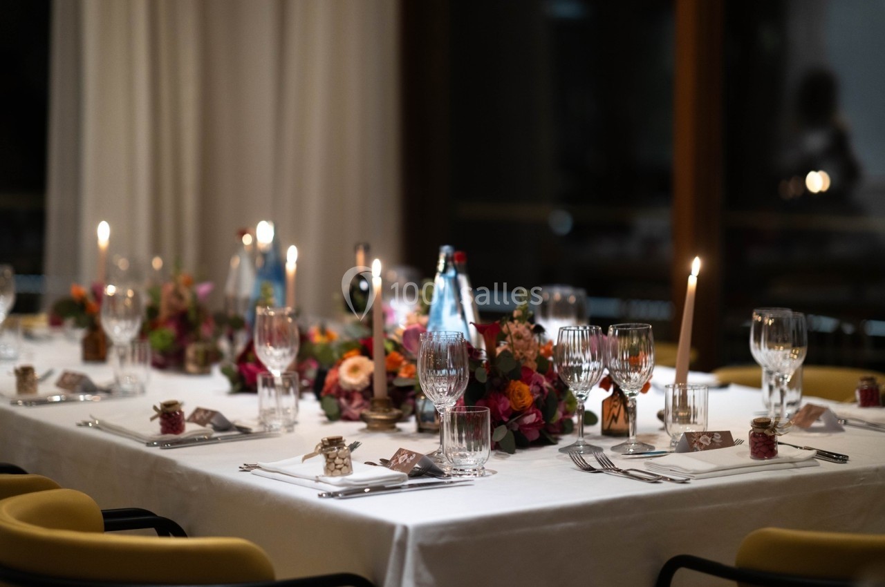 Table élégamment dressée avec nappes blanches, verres, bougies allumées et décorations florales colorées.